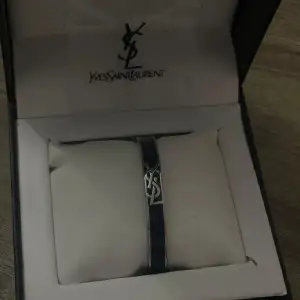 Snyggt och stilrent armband från Yves Saint Laurent i en elegant svart färg. Armbandet har en ikonisk YSL-logga i silver som ger en lyxig touch. Perfekt accessoar för både vardag och fest. Kommer i originalförpackning.