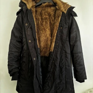 Svart vinterjacka med fuskpäls - Säljer en svart vinterjacka med fuskpälsfoder och huva. Jackan har dragkedja och knappar framtill, samt praktiska fickor med dragkedjor. Perfekt för kalla vinterdagar! 🖤