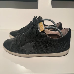Svarta sneakers från Golden Goose - Snygga svarta sneakers från Golden Goose i modellen superstar skorna är storlek 37 och innermåttet är ca 23,5cm . De har en cool, lite sliten look och är perfekta för vardagsbruk. Skorna har en klassisk snörning och en bekväm sula. Perfekta för dig som vill ha en stilren men ändå unik look. Skorna är väl använda därav priset, ingen originallåda medföljer. Tveka inte att skriva vid minsta fundering. 👌🥂👊priset är inte hugget i sten.