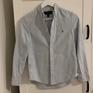 Randig skjorta från Ralph Lauren - Säljer en stilren och klassisk skjorta från Ralph Lauren i blå och vit randigt mönster. Skjortan har långa ärmar och knappar framtill. Den är broderad med den ikoniska loggan på bröstet. Perfekt för både vardag och fest! 🕺