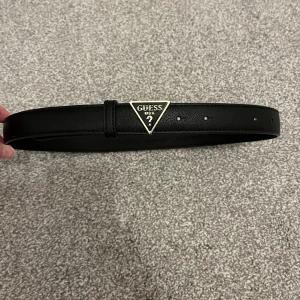 Svart bälte från Guess - Säljer ett stilrent svart bälte från Guess i skinn imitation. Det har en snygg triangel-logga i guld som ger en lyxig touch. Perfekt för att piffa upp vilken outfit som helst, oavsett om det är till vardags eller fest. Bältet är i nytt skick och passar perfekt till sommarens alla tillfällen.