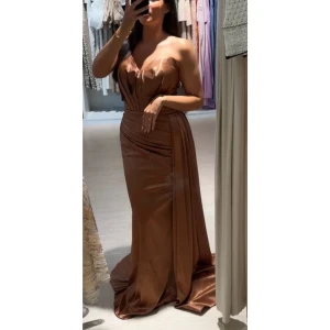 Bronze/brun axelbandslös i satin långklänning - Elegant bronze/brun axelbandslös långklänning i satin med draperad design och slits. Perfekt för festliga tillfällen som bröllop och förlovning  med en smickrande passform och vacker glans.