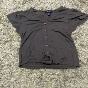 Mörkgrön topp från New Look - Säljer en mörkgrön topp från New Look i storlek 12/13 år. Toppen har korta ärmar och knappar framtill, samt en knytning vid nederkanten för en avslappnad look. Perfekt för vardagsbruk!