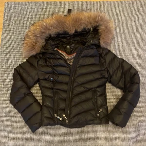 Svart dunjacka med pälsdetaljer från Hollies - Säljer en snygg svart dunjacka från Hollies med en fluffig pälsdetalj på huvan. Jackan har dragkedja och två fickor framtill. Insidan har ett elegant mönster i grått och svart. Går även att ta bort all päls som finns på jackan om man föredrar det. Kan diskutera om pris❤️