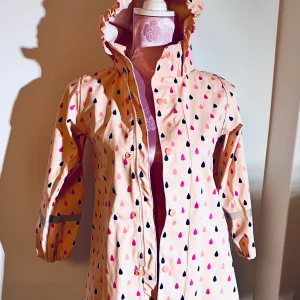 Regnkappa Reima - Rosa regnkappa med droppmönster. Reflexer på ärmarna, bak och på kapuschong. Kapuschong är avtagbar. Stängs med både blixtlås och tryckknappar.