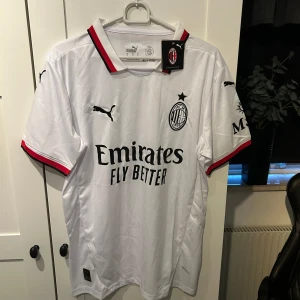 Vit AC Milan fotbollströja från Puma - Säljer en vit AC Milan fotbollströja från Puma i nytt skick. Tröjan har korta ärmar och snygga röda och svarta detaljer vid krage och ärmslut. Den är gjord i ett lätt och bekvämt material, perfekt för både träning och match. Stor logga och sponsortryck på framsidan. Passa på att köpa denna stilrena tröja!