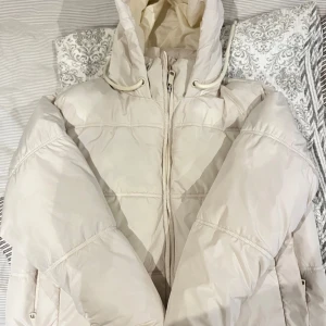 Beige pufferjacka med huva - Säljer en snygg beige/vit pufferjacka från Zara i storlek S. Den har en praktisk jättestor huva och dragkedja framtill. Perfekt för kyliga dagar och håller dig varm och bekväm. Jackan är i bra skick men har en liten fläck som jag tror jag går bort om man tvättar den ☺️ 