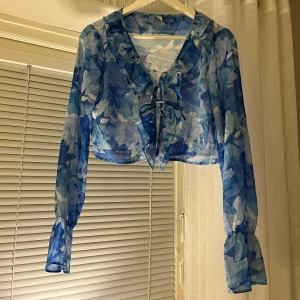 Blå blommig blus med volangdetaljer - Säljer en superfin blå blommig blus med volangdetaljer framtill. Blusen har långa ärmar och är perfekt för både vår och sommar. Den är lätt och luftig, vilket gör den idealisk för varmare dagar. Passar perfekt till både jeans och kjol!