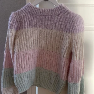 Randig stickad tröja från Vero Moda - Superfin stickad tröja från Vero Moda i pastellfärger. Den har breda ränder i lila, beige, rosa och mintgrön. Perfekt för kyliga dagar med sin mysiga och varma känsla. Tröjan har långa ärmar och en ribbad kant vid halsen.