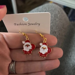 Julörhängen med tomtemotiv - Söta örhängen med tomtemotiv i rött och vitt, perfekta för julens festligheter. De har guldfärgade krokar och är lätta att bära. Perfekt accessoar för att sprida julstämning! 🎅✨