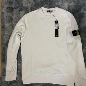 Vit tröja från Stone Island - Säljer en snygg vit tröja från Stone Island i nytt skick. Den har en klassisk design med rund hals och långa ärmar. Tröjan är i bomull och har det ikoniska Stone Island-märket på ärmen. Perfekt för både vardag och fest!