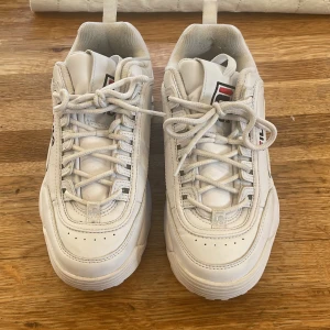 Vita sneakers från Fila - Säljer ett par vita Fila sneakers i mycket bra skick. De har en chunky sula och klassisk snörning. Perfekta för både vardag och träning. Storlek 38 Fila-loggan syns tydligt på sidan och baksidan av skorna.