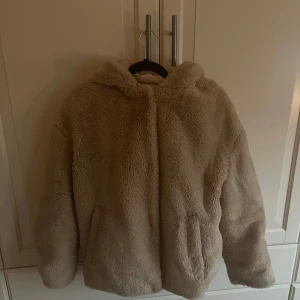 Beige teddyjacka med huva - Mysig beige teddyjacka med huva och knappar. Den har ett hål i och en knapp som sitter löst
