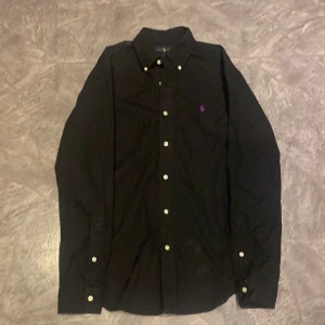 Svart skjorta från Ralph Lauren - Snygg svart skjorta från Ralph Lauren i slim fit. Skjortan har långa ärmar och knappar framtill. Den är dekorerad med en liten broderad logotyp i lila på bröstet. Perfekt för en stilren look.