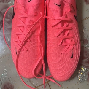 Rosa Nike fotbollsskor - Snygga rosa Nike fotbollsskor med svart logga och snörning. Skorna har en modern design med Nike Gripknit-teknologi för bättre grepp och kontroll på planen. Perfekta för den som vill sticka ut på fotbollsplanen.