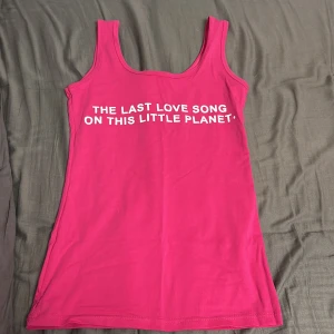 Rosa linnetopp med text - Säljer en rosa y2k linnetopp med texten 'THE LAST LOVE SONG ON THIS LITTLE PLANET'. Toppen har en enkel design med rund halsringning och ärmlös stil. Perfekt för en avslappnad look. Köptes utomlands så den har inget märke. Säljs för jag har ingen användning av den. 