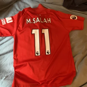 Röd Liverpool fotbollströja med M. Salah 11 - Säljer en röd Liverpool fotbollströja från New Balance med M. Salah.  Den är storlek 152. Perfekt för fans av laget!