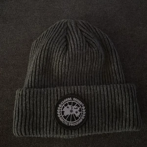 Grå ribbad mössa från Canada Goose - Snygg grå ribbad mössa från Canada Goose med ett broderat märke framtill. Perfekt för kyliga dagar och ger en stilren look. Passar bra till både vardag och utomhusaktiviteter.