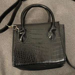 Svart axelväska med mönster - Snygg svart axelväska med krokodilmönster och avtagbar axelrem. Väskan har två handtag och en stilren design som passar perfekt för en trendig look. Perfekt för att bära dina dagliga nödvändigheter med stil.