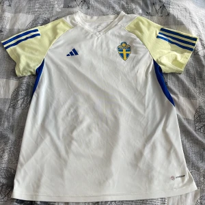 Vit fotbollströja med gula och blå detaljer från Adidas - Säljer en vit fotbollströja från Adidas med gula ärmar och blå ränder. Tröjan har korta ärmar och ett tryckt emblem på bröstet. Perfekt för fotbollsfans!