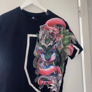 Svart t-shirt med draktryck från Horspist - Horspist svart t-shirt med ett stort, färgglatt draktryck på framsidan. T-shirten har korta ärmar och en rund halsringning, storlek L men sitter som Medium.