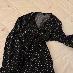 Svart prickig klänning - Snygg svart klänning med vita prickar och v-ringad design. Klänningen har långa ärmar och en smickrande passform med knyt i midjan. Perfekt för en stilren look.