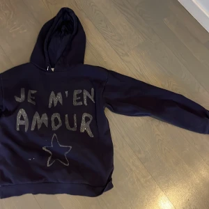 Mörkblå hoodie från Gina tricot - Säljer en mörkblå hoodie från Mini Rodini med texten 'JE M'EN AMOUR' i glittriga bokstäver och en stjärna på framsidan. Perfekt för en avslappnad stil med en touch av glitter. 🩵