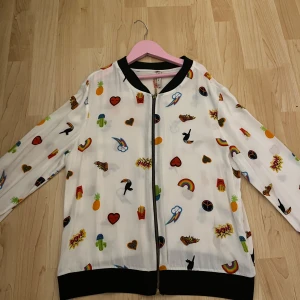 Vit bomberjacka med färgglada mönster från Danque - Säljer en cool vit bomberjacka från Danque med färgglada mönster som hjärtan, regnbågar och kaktusar. Jackan har en dragkedja framtill och svarta muddar vid ärmslut och nederkant. Perfekt för att liva upp din outfit! 🌈