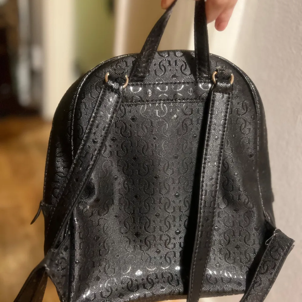 Snygg svart liten ryggsäck från Guess med ett subtilt mönster och guldfärgade detaljer. Väskan har en praktisk dragkedja och justerbara axelremmar. Perfekt för att bära dina dagliga nödvändigheter med stil.. Laukut & Käsilaukut.