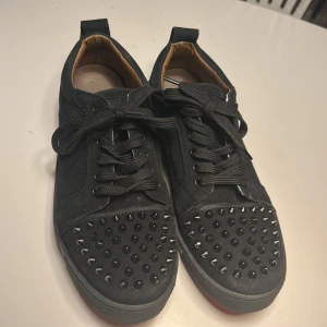Svarta sneakers med nitar från Christian Louboutin - Snygga svarta sneakers från Christian Louboutin med nitar på tån och den ikoniska röda sulan. Skorna har snörning och är tillverkade i mocka. Perfekta för att ge din stil en edgy touch. Storlek 44 men passar 43 med och det är två nitar som har lossnat. Kom för mer bilder.pris kan diskuteras 