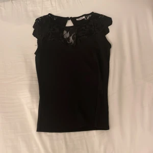 Svart topp med spetsdetaljer från Only - Snygg svart topp från Only med spetsdetaljer vid halsringningen och axlarna. Toppen har en elegant nyckelhålsöppning bak och är ärmlös, vilket gör den perfekt för både vardag och fest.❤️