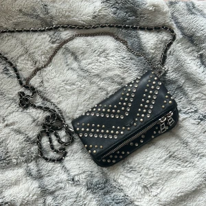 Svart axelväska med nitar från Zadig & Voltaire - Snygg svart axelväska i skinn från Zadig & Voltaire. Väskan är dekorerad med silver- och guldfärgade nitar i ett coolt mönster. Den har en kedjerem och en praktisk dragkedja framtill. Perfekt för att ge din outfit en edgy touch. Den är lite rostig på nitarna och den främre dragkedjan men funkar som en ny, den här väskan är även limited edition och då är svår att få☺️