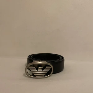 Svart skärp med silverfärgat spänne - Snyggt svart skärp från Emporio Armani med ett stilrent silverfärgat spänne. Perfekt för att ge en elegant touch till din outfit. Skärpet är gjort av skinn och har en klassisk design som passar till många olika stilar.