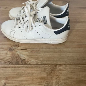 Vita Adidas Stan Smith sneakers - Säljer ett par klassiska vita Adidas Stan Smith sneakers med marinblå detaljer. Skorna har snörning och den ikoniska loggan på hälen. Perfekta för en stilren look. Sparsamt använda, säljer då de tyvärr är för små för mig. De är i storlek 38,2/3💖nypris är 900