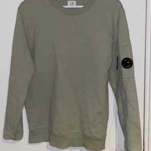 Olivgrön tröja från C.P. Company - Säljer en stilren olivgrön tröja från C.P. Company. Tröjan har långa ärmar och en rund halsringning. På ärmen finns en unik detalj i form av en svart rund emblem. Perfekt för en avslappnad och trendig look.  Skriv vid frågor eller bilder 