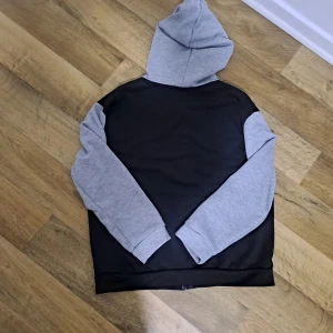 Svart och grå hoodie med dragkedja  - Snygg hoodie med svart kropp och grå ärmar samt huva. Perfekt för en avslappnad stil. Den har en klassisk design med långa ärmar och en bekväm passform.