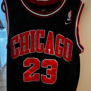 Svart Chicago Bulls basketlinne från Nike - Säljer ett svart basketlinne från Nike med Chicago Bulls tema. Linnet har nummer 23 och texten 'CHICAGO' i rött med vita konturer. Perfekt för basketfans och sportentusiaster! Storlek M