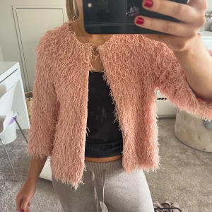 Rosa fluffig jacka - Säljer en supermjuk och fluffig rosa kofta med trekvartsärm. Perfekt för att ge din outfit en unik touch. Koftan har en öppen design med en knapp högst upp. 