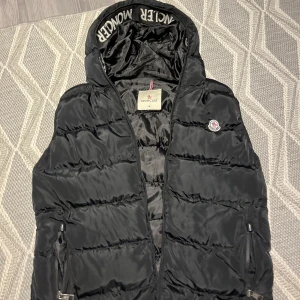 Svart dunväst från Moncler - Snygg svart dunväst från Moncler med dragkedja och huva. Perfekt för kyligare dagar och ger en stilren look. Moncler-logga på bröstet och märkesdetaljer på insidan av huvan.