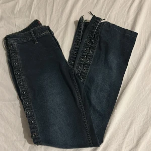 Lågmidjade Gazoz Jeans - Snygga mörkblå jeans från Gazoz Jeans med unik snörning längs sidorna. De har en klassisk femficksdesign och knappgylf. Perfekta för dig som vill ha en edgy stil.