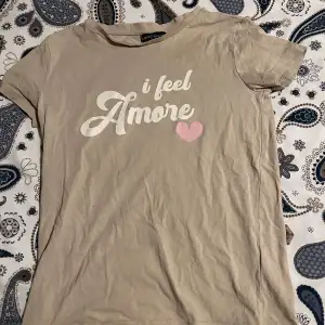 En vanlig beige t-Shirt med text. Texten är ”I Feel Amore”. Kommer inte till användning här hemma, bra skick.