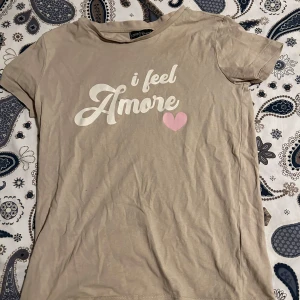 Beige t-shirt  - En vanlig beige t-Shirt med text. Texten är ”I Feel Amore”. Kommer inte till användning här hemma, bra skick.