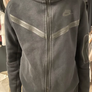 Svart hoodie från Nike - Säljer en svart hoodie från Nike tec i bra skick. Den har en dragkedja framtill och en liten ficka med dragkedja på ärmen. Den är superbekväm med normal passform. Nike-loggan syns diskret på bröstet. Den är ca ett år gammal och i storlek 147-158