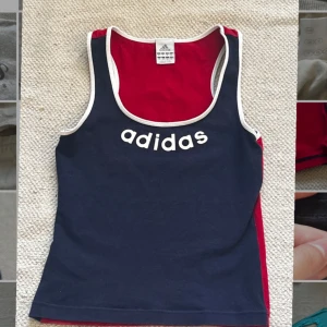 Blå och röd linnetopp från Adidas - Säljer en snygg och sportig blå och röd linnetopp från Adidas. Den har ett klassiskt Adidas-tryck på framsidan och en stor siffra '3' på baksidan. Den är sydd på sidan då den var för stor, inget som påverkar tröjan den är bara smalare. Skriv frågor❤️💙