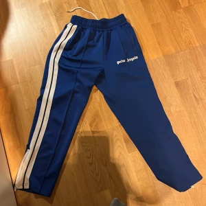 Blå byxor från Palm Angels - Säljer ett par snygga blå byxor från Palm Angels. De har en vit rand längs sidorna och en elastisk midja för extra komfort. Perfekta för en avslappnad stil eller träning. Byxorna har en normal passform och är gjorda i ett mjukt material.