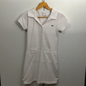 Vit klänning från Lacoste - Säljer en snygg vit klänning från Lacoste med korta ärmar och en klassisk krage. Klänningen har en stilren design med två praktiska fickor framtill och en diskret Lacoste-logga på bröstet. Perfekt för en avslappnad sommarlook eller en dag på tennisbanan!