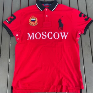 Röd pikétröja från Ralph Lauren - Säljer en snygg röd pikétröja från Ralph Lauren med texten 'MOSCOW' broderad på framsidan. Tröjan har en klassisk krage med vita och svarta detaljer och en stor svart siffra '3' på ryggen. Den har korta ärmar och är perfekt för en sportig look.