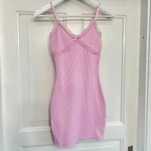 Rosa randig klänning med spetsdetaljer - Säljer min rosa klänning med spetsdetaljer vid bysten. Den har smala justerbara axelband och en liten rosett framtill. Klänningen är i ett mjukt och stretchigt material. Storlek XS från H&M. Nyskick. Aldrig använd.