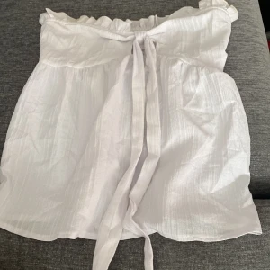 Vit axelbandslös topp med knytband - Säljer en superfin vit axelbandslös topp med knytband framtill. Den har en smockad överdel som ger en bekväm passform och volangdetaljer längs kanten. Perfekt för sommardagar eller en festlig kväll! Aldrig använd!!
