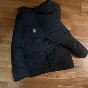 Svart dunjacka från trapstar - Snygg svart dunjacka från Trapstar  med huva och dragkedja. Jackan har ett broderat märke på ryggen och praktiska fickor framtill. Perfekt för kyliga dagar.